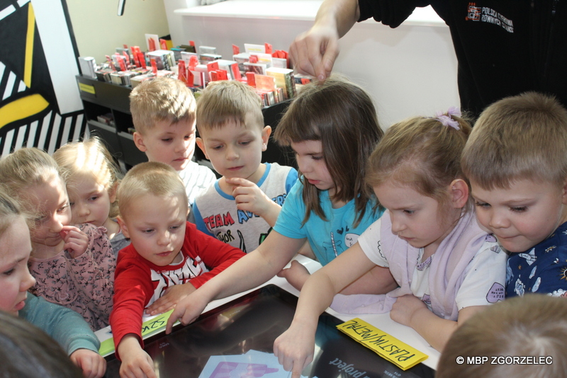 W pomieszczeniu biblioteki, grupa przedszkolaków dotyka ekran multimedialny i sortuje wyświetlane karty ilustrujące odpady.