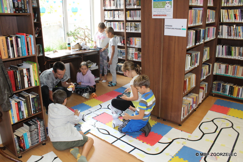 W pomieszczeniu biblioteki po piankowej macie jeżdżą zaprogramowane roboty. Obok siedzą dzieci i programują trasy dla robotów z użyciem białych arkuszy z nadrukowaną czarną linią..