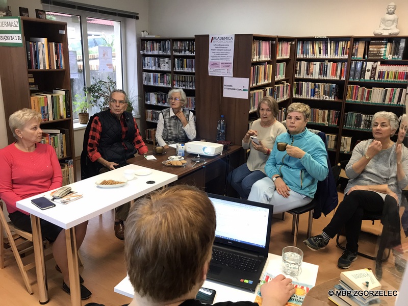 Sześcioro seniorów siedzi w pomieszczeniu biblioteki podczas spotkania. Na stołach pośrodku znajdują się ciastka na talerzykach i butelka wody mineralnej. Na pierwszym planie widać tył głowy prowadzącego i laptopa. 