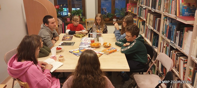 W pomieszczeniu czytelni Oddziału Dla Dzieci i Młodzieży MBP w Zgorzelcu siedzi i dyskutuje przy stołach 9 osób.