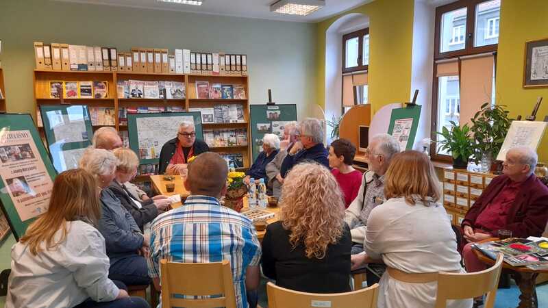 Grupa osób siedzi i rozmawia przy stole w bibliotecznej czytelni. Na stole znajdują się filiżanki kawy i butelki z wodą.