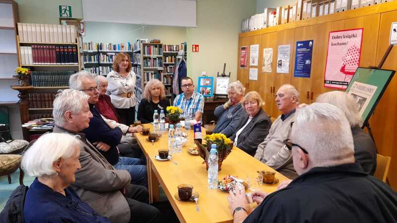 Grupa osób siedzi i rozmawia przy stole w bibliotecznej czytelni. Na stole znajdują się filiżanki kawy i butelki z wodą.