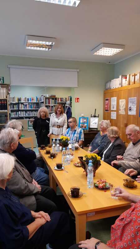 Grupa czytelników jest witana przez prowadzące podczas spotkania w bibliotecznej czytelni. Uczestnicy siedzą przy stole, na którym znajdują się filiżanki z kawą, butelki z wodą i kwiaty.