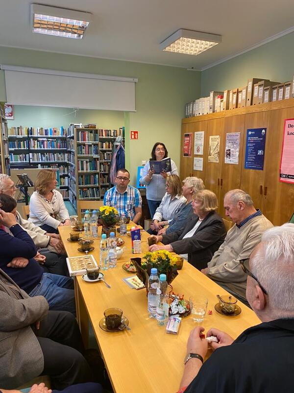 Kobieta stoi i czyta z książki podczas spotkania w bibliotecznej czytelni. Pozostali uczestnicy słuchają, siedząc przy stole, na którym znajdują się filiżanki z kawą, butelki z wodą i kwiaty.