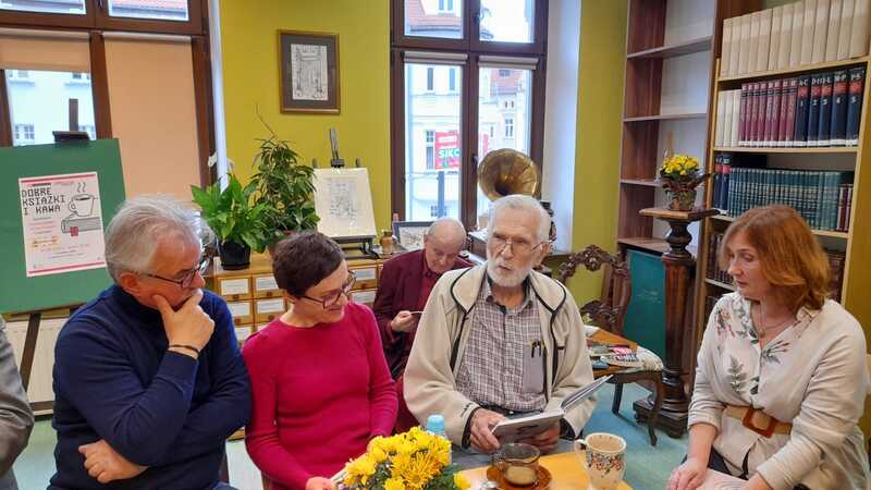 Grupa osób rozmawia przy książce w bibliotecznej czytelni. Siedzą przy stole, na którym znajdują się filiżanki z kawą i kwiaty.