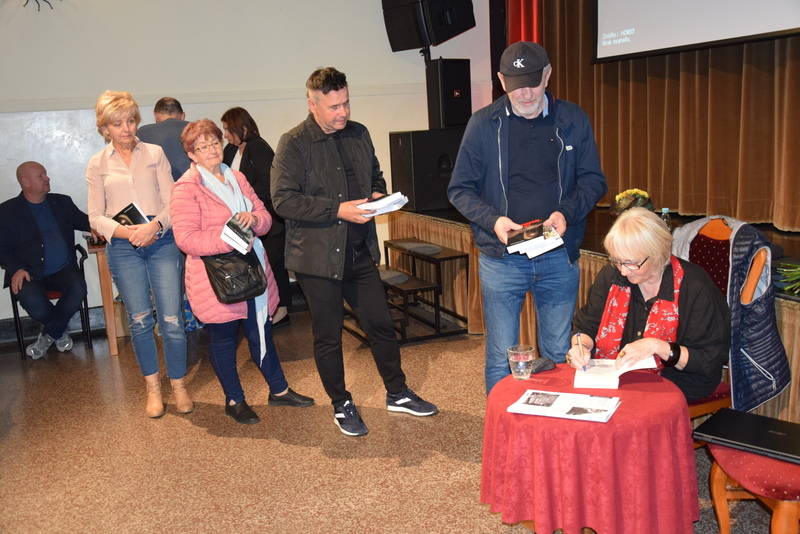 Autorka podpisuje książki po spotkaniu, siedząc przy czerwonym stoliku. Stoi do niej długa kolejka czytelników z książkami w dłoniach.