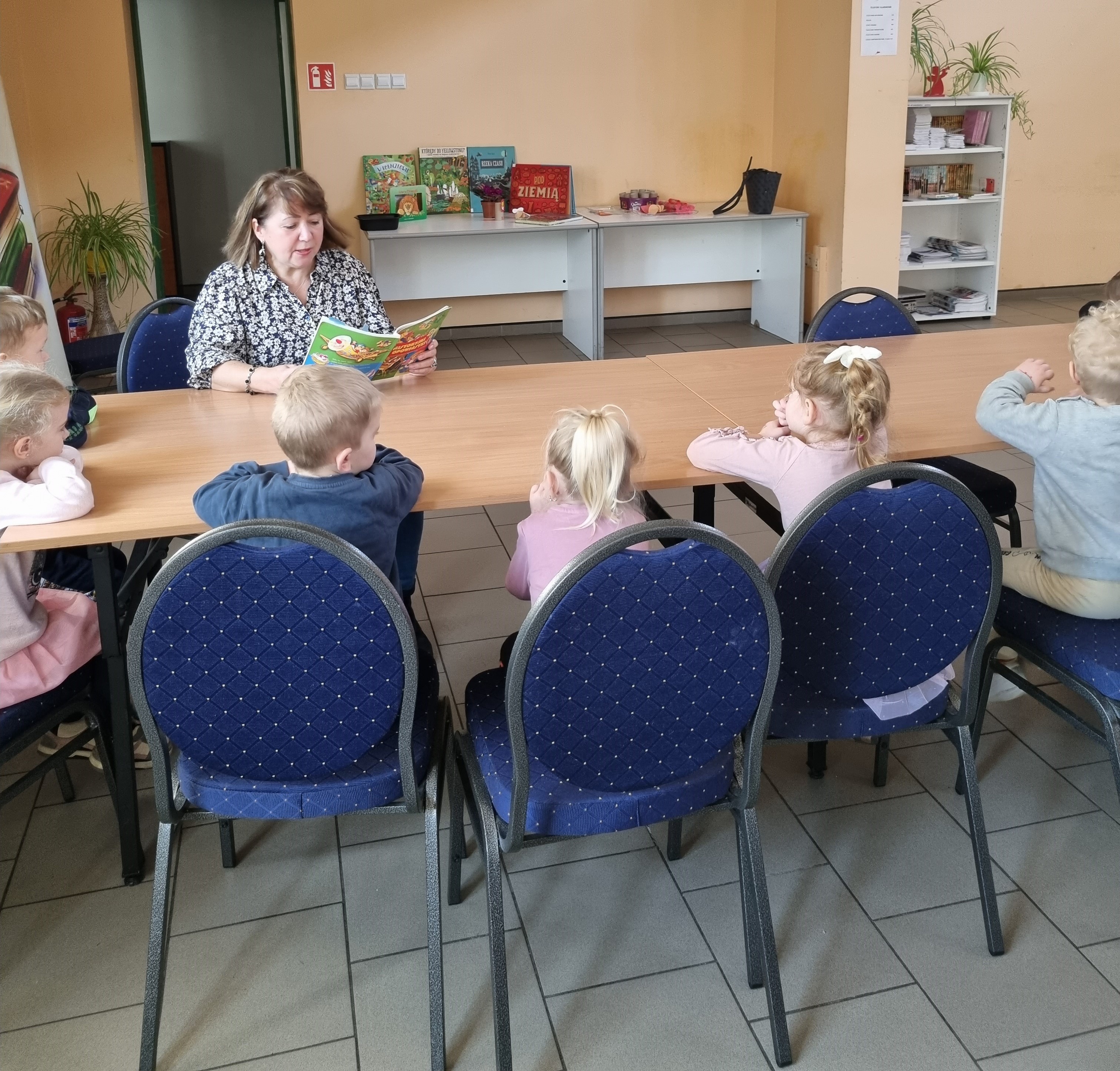 Bibliotekarka siedzi przy długim stole z jasnego drewna naprzeciwko dzieci w wieku przedszkolnym i czyta na głos z książki dla dzieci.