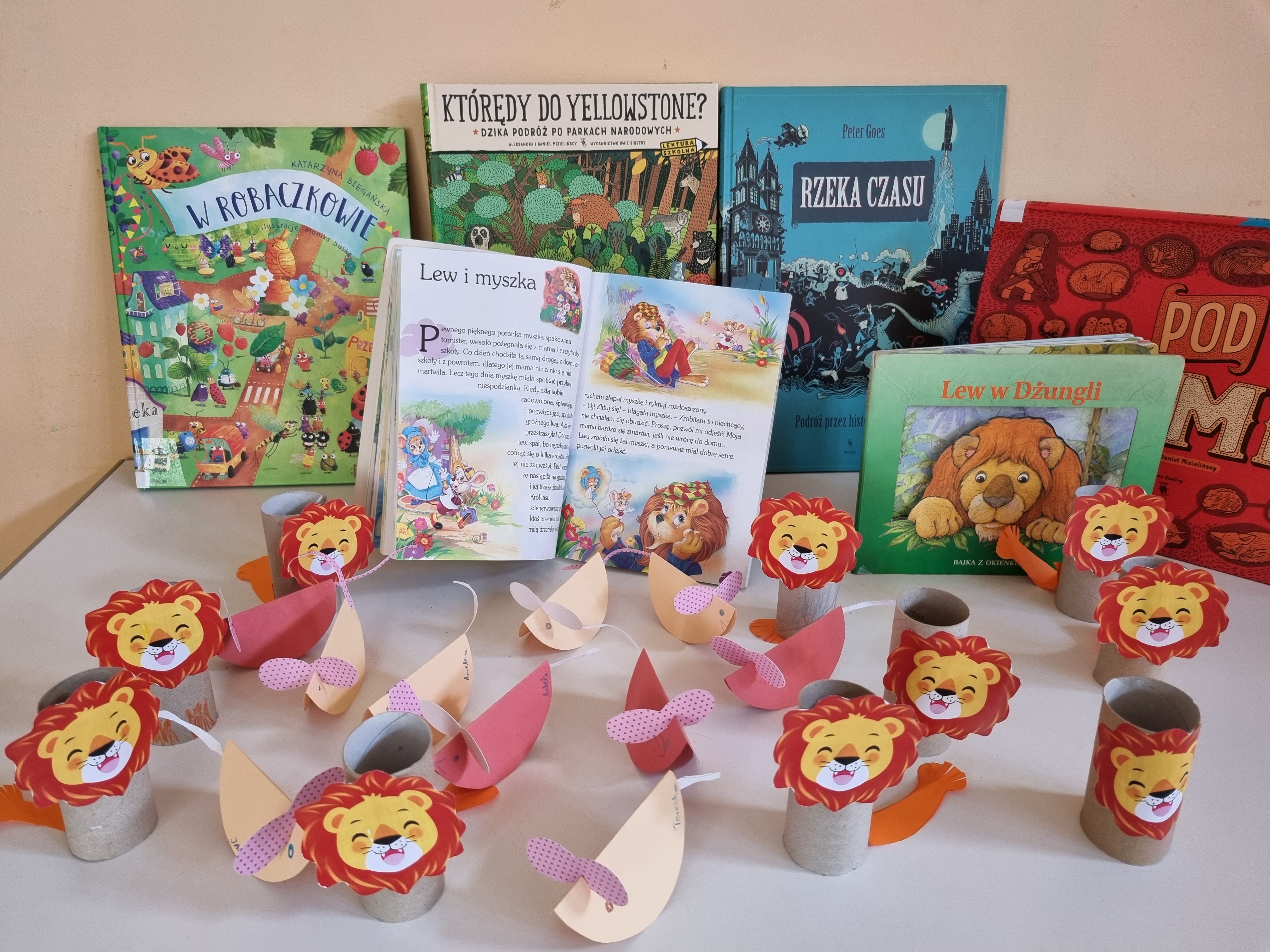 Na jasnym stole leżą papierowe prace plastyczne przedstawiające lwy, wykonane przez dzieci. Za nimi ustawione są kolowe książki dla dzieci, niektóre z nich mówią o lwach.