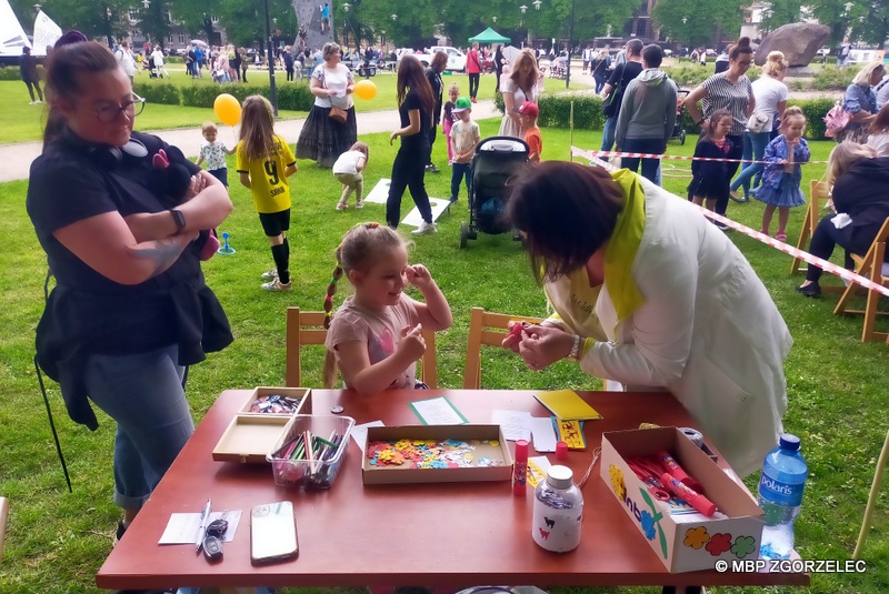 Przy stoliku dziewczynka przygotowuje laurkę i zakładkę okolicznościową z okazji Dnia Matki. Obok stoją dwie kobiety a z tyłu grupka dzieci bierze udział w zabawach animacyjnych.