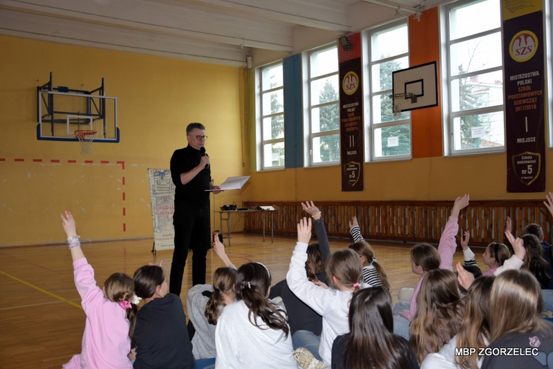 W pomieszczeniu sali gimnastycznej Szkoły Podstawowej nr 5 im. Marii Skłodowskiej-Curie, pan Marek Michalak opowiada czyta dzieciom fragment swojej książki..