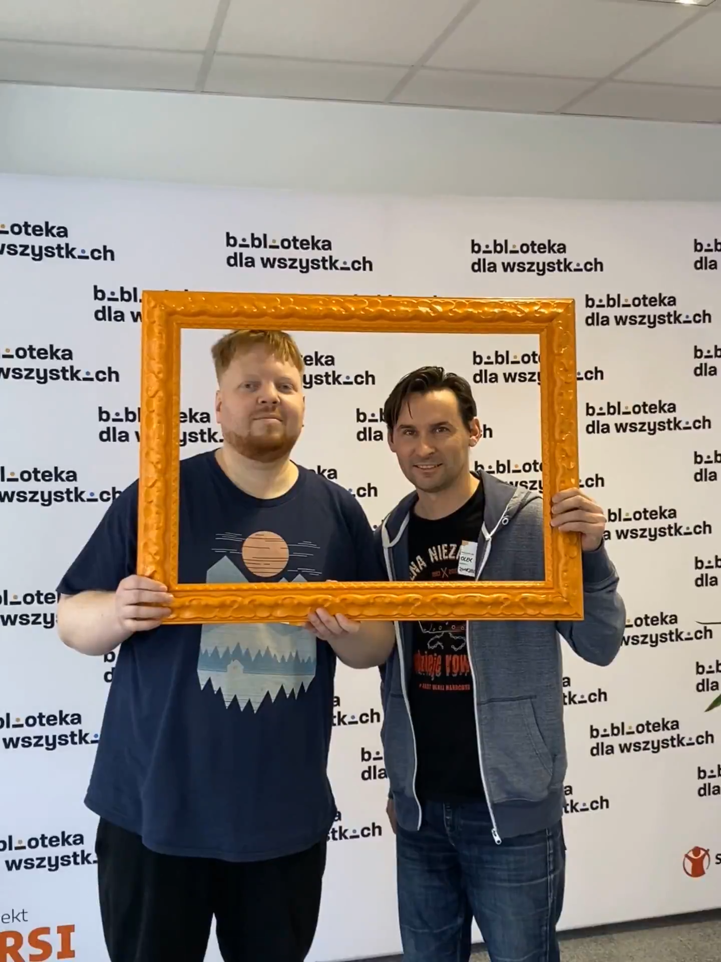 W pomieszczeniu, na tle ściany z napisami "Biblioteka dla wszystkich" stoi dwoje bibliotekarzy, którzy trzymają pomarańczową ramkę na obraz i pozuje do zdjęcia.