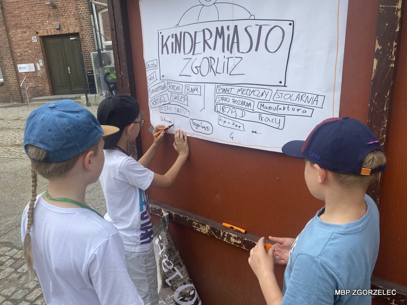Troje dzieci zapisuje duży plakat zatytułowany 'Kindermiasto Zgorlitz', przedstawiający schemat miasta z oznaczeniami instytucji takich jak urząd, bank, sklep i szpital. Plakat przymocowany jest do drewnianej tablicy, a dzieci używają czarnych markerów.