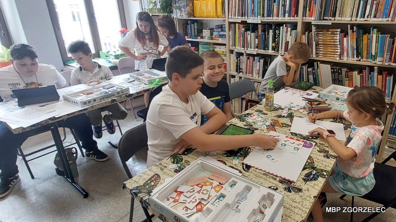 Dzieci z wykorzystaniem zestawów planszowych Scottie Go Edu programują aniowane postacie w aplikacji na tabletach przy stołach w bibliotece.