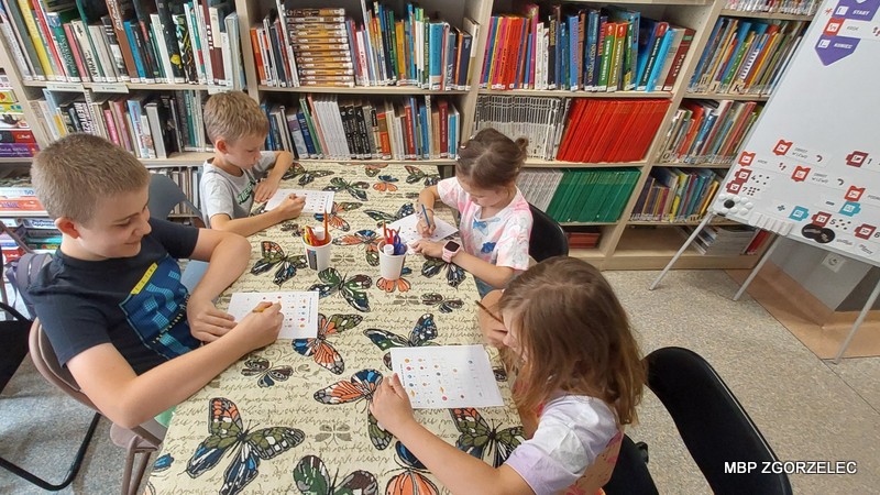 Czwórka dzieci siedzących przy stole w bibliotece. Dzieci pracują z kartami pracy i kolorowymi kredkami. Stół przykryty jest obrusem z motywem motyli, a w tle widoczne są regały pełne książek oraz tablica z planszami do programowania. 