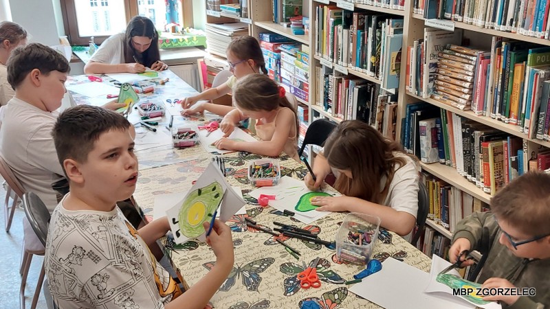 Dzieci rysują, wycinają i tworzą prace plastyczne przy stole w bibliotece.