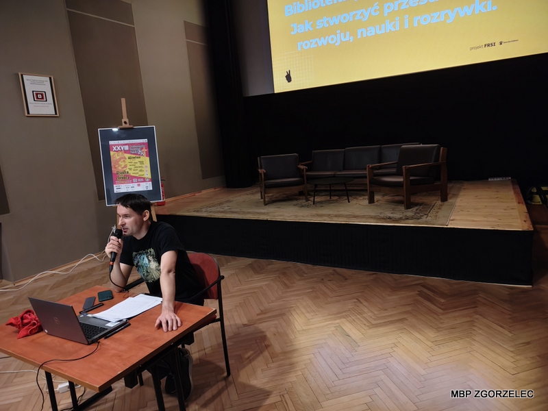 W sali kinowej Miejskiego Domu Kultury w Zgorzelcu bibliotekarz siedzi przy stole z laptopem i mikrofonem, prowadzi wystąpienie. W tle znajduje się scena z krzesłami oraz duży ekran z tekstem w języku polskim: „Biblioteki... Jak stworzyć przestrzeń rozwoju, nauki i rozrywki”. W sali kinowej Miejskiego Domu Kultury w Zgorzelcu bibliotekarz siedzi przy stole z laptopem i mikrofonem, prowadzi wystąpienie. W tle znajduje się scena z krzesłami oraz duży ekran z tekstem w języku polskim: „Biblioteki... Jak stworzyć przestrzeń rozwoju, nauki i rozrywki”.