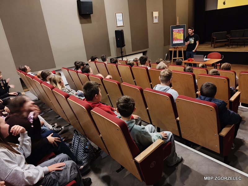 Grupa dzieci siedzi w sali kinowej Miejskiego Domu Kultury w Zgorzelcu. Dzieci zajmują czerwone fotele ustawione w rzędach i z uwagą patrzą przed siebie na bibliotekerza, który przedstawia prezentację dot. nowej przestrzeni bibliotecznej dla młodzieży w Zgorzelcu. Grupa dzieci siedzi w sali kinowej Miejskiego Domu Kultury w Zgorzelcu. Dzieci zajmują czerwone fotele ustawione w rzędach i z uwagą patrzą przed siebie na bibliotekerza, który przedstawia prezentację dot. nowej przestrzeni bibliotecznej dla młodzieży w Zgorzelcu.
