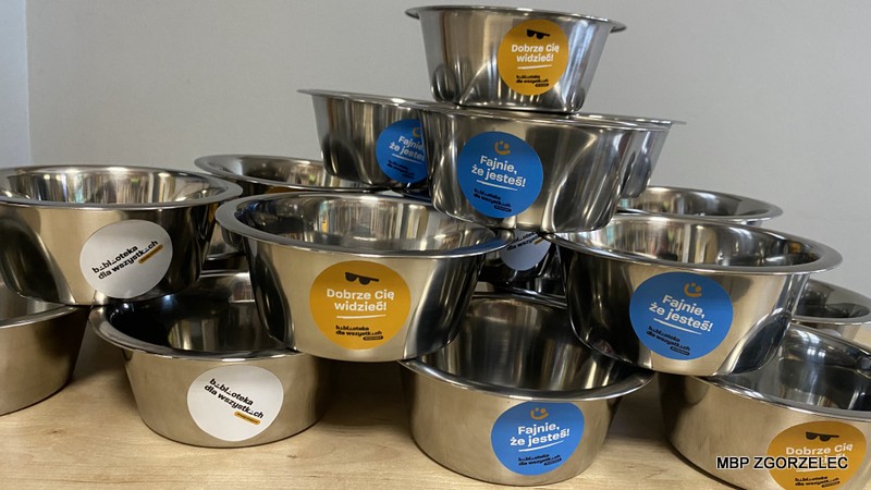 Stos metalowych misek dla zwierząt ustawionych w piramidę, każda z kolorową naklejką z hasłami „Dobrze Cię widzieć!” i „Fajnie, że jesteś!”. Miski są częścią akcji „Biblioteka dla wszystkich istot”.