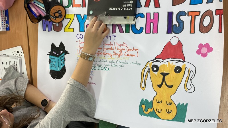 Osoba rysująca kolorowy plakat z napisem „Biblioteka dla wszystkich istot”, ozdobiony ilustracjami kota z książką i psa w czapce Mikołaja.