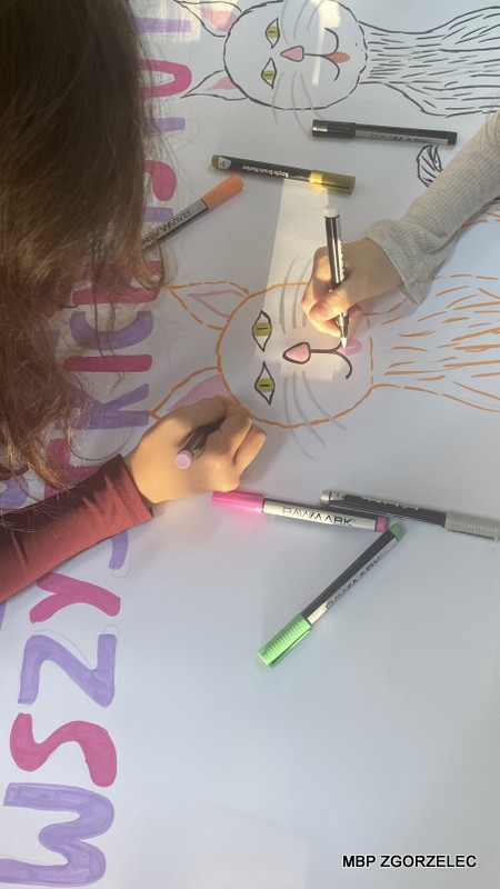 Zbliżenie na dłonie osób rysujących kolorowego kota na dużym plakacie markerami w różnych kolorach.