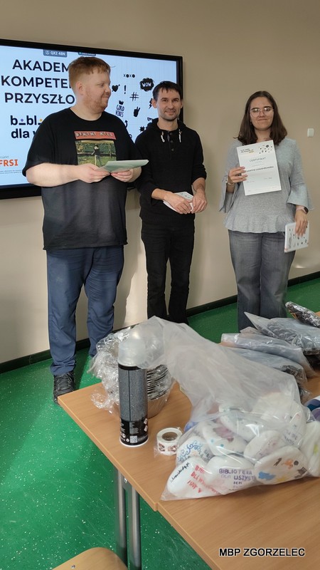 Trzy osoby stojące w sali lekcyjnej przy stole z materiałami na rzecz schroniska. Jedna osoba trzyma notatniki, druga trzyma certyfikaty w ramach programu „Akademia Kompetencji Przyszłości” realizowanego przez bibliotekę i przekazuje je absolwentce programu.