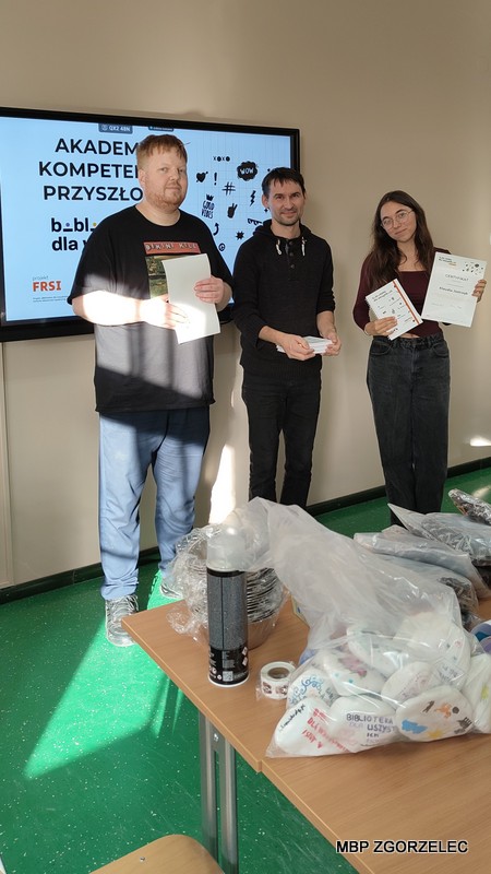 Trzy osoby stojące w sali lekcyjnej przy stole z materiałami na rzecz schroniska. Jedna osoba trzyma notatniki, druga trzyma certyfikaty w ramach programu „Akademia Kompetencji Przyszłości” realizowanego przez bibliotekę i przekazuje je absolwentce programu.