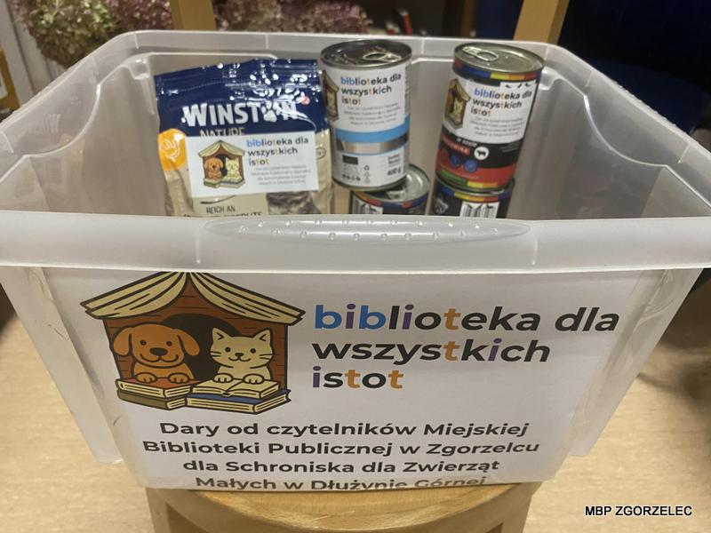 Plastikowy pojemnik z naklejką „Biblioteka dla wszystkich istot” wypełniony puszkami karmy dla psów i kotów oraz paczką przysmaków, przygotowany do przekazania schronisku.