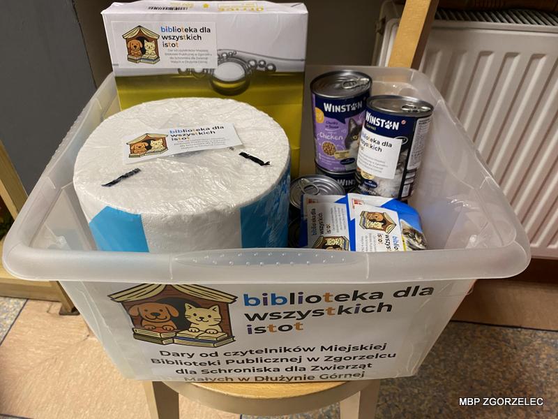 Plastikowy pojemnik z naklejką „Biblioteka dla wszystkich istot” zawierający dary od czytelników Miejskiej Biblioteki Publicznej w Zgorzelcu dla schroniska w Dłużynie Górnej. W środku widoczne rolki papieru, puszki karmy dla psów i kotów oraz akcesoria.