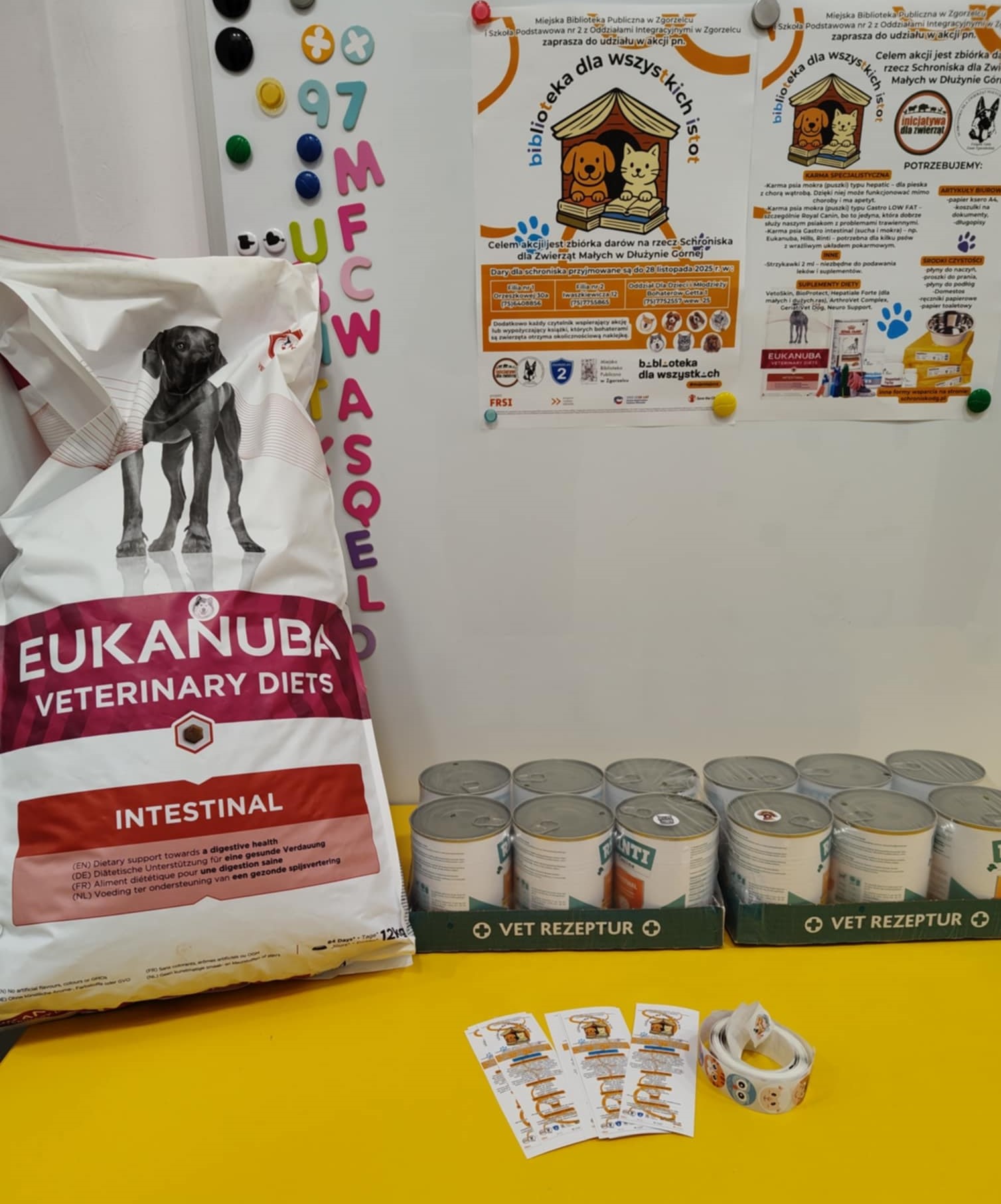 Duża torba karmy Eukanuba Veterinary Diets oraz kilkanaście puszek karmy weterynaryjnej ustawionych na żółtym stole. Obok leżą ulotki akcji „Biblioteka dla wszystkich istot”, a w tle widoczna tablica z plakatami informacyjnymi.