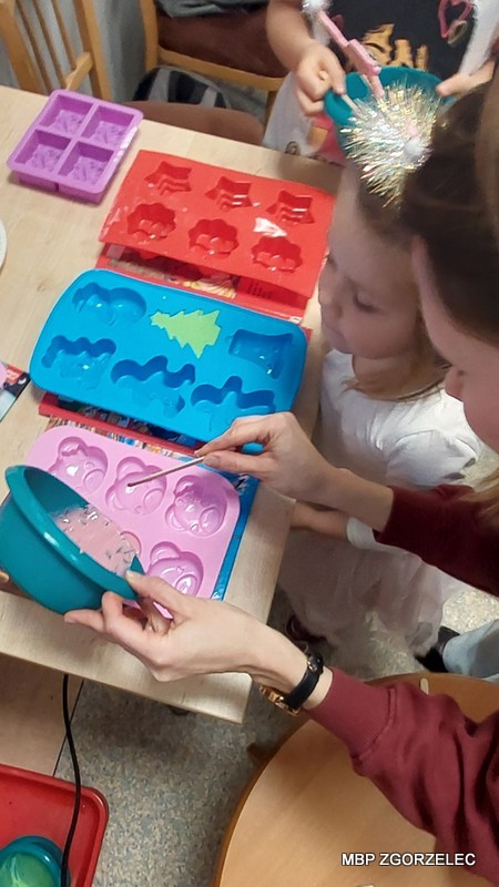 Warsztaty tworzenia mydełek – na stole foremki silikonowe w różnych kształtach, bibliotekarka pomaga dziecku w przygotowaniu masy.