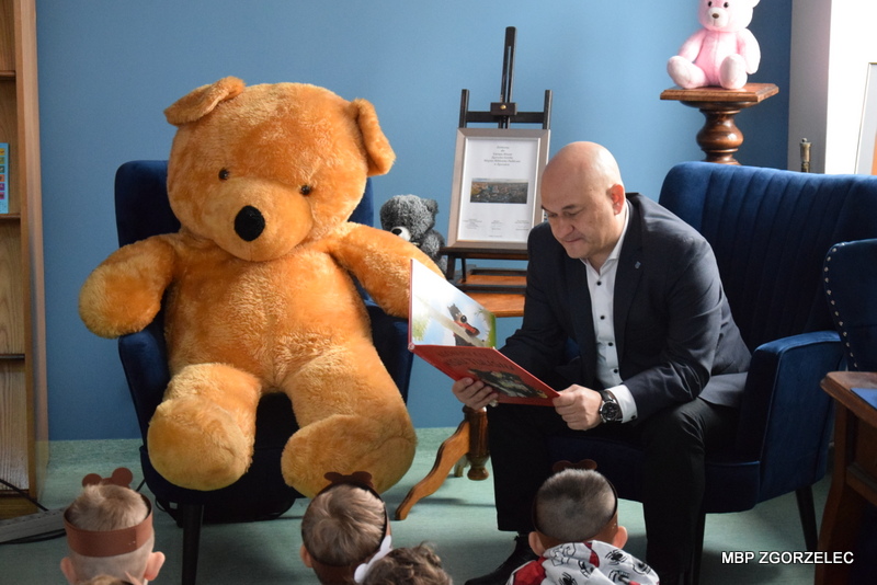 Czytanie książki dzieciom w towarzystwie pluszowego misia – Pan burmistrz Miasta Zgorzelec - Rafał Gronicz siedzi obok dużego misia i czyta książkę grupie dzieci, które uważnie słuchają. W tle widoczne są półki z książkami i dekoracje.