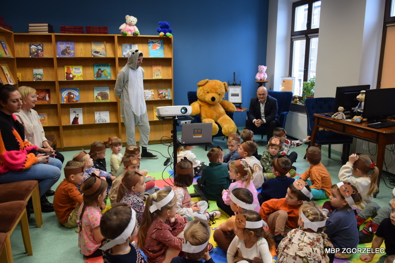 W czytelni biblioteki dzieci uczestniczą w zajęciach, obok nich stoi bibliotekarz w kostiumie koali, obok na niebieskim fotelu siedzi Pan burmistrz Rafał Gronicz i pluszowy miś. W tle są widoczne półki z książkami.