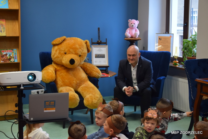 Przestrzeń czytelni z niebieskimi fotelami, dużym pluszowym misiem siedzącym na jednym z nich oraz z Panem burmistrzem - Rafałem Groniczem siedzącym na drugim fotelu. Na zdjęci widać także projektor z grupą dzieci w opaskach z motywem misia, uczestniczących w zajęciach literackich.