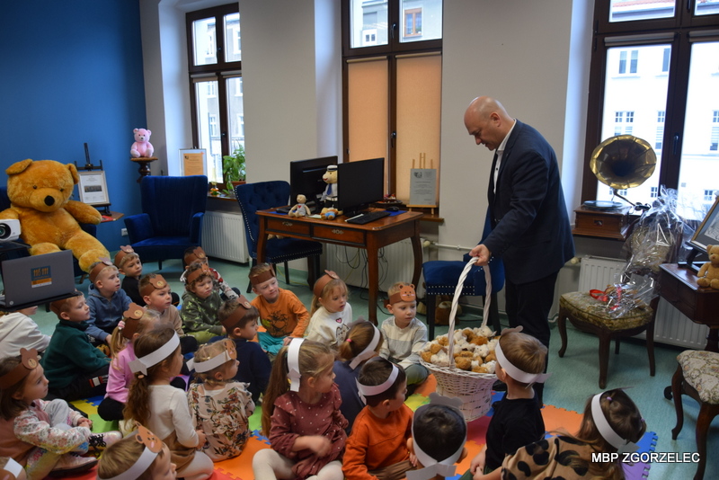 Wręczanie pluszowych misiów dzieciom –Pan burmistrz Rafał Gronicz rozdaje dzieciom maskotki z dużego kosza, dzieci siedzą na kolorowych matach w czytelni.