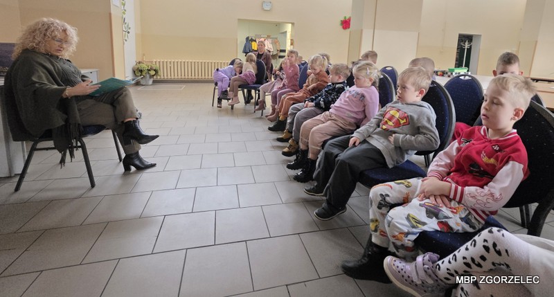 Czytanie bajek w sali Miejskiego Klubu Seniora – dzieci siedzą w rzędzie na krzesłach, a Pani dyrektor biblioteki - Krystyna Radzięta czyta im książkę.