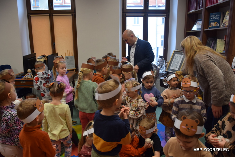 Wręczanie pluszowych misiów dzieciom –Pan burmistrz Rafał Gronicz rozdaje dzieciom maskotki z dużego kosza, dzieci siedzą na kolorowych matach w czytelni.