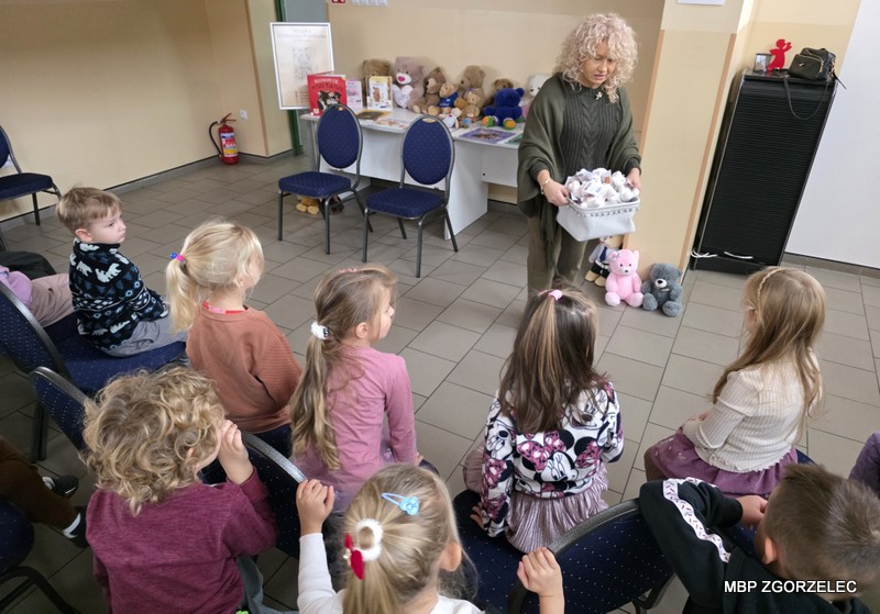 Dzieci siedzą na krzesłach, a Pani dyrektor biblioteki - Krystyna Radzięta rozdaje dzieciom z koszyka pluszowe misie. Na stole w tle znajdują się książki i maskotki.