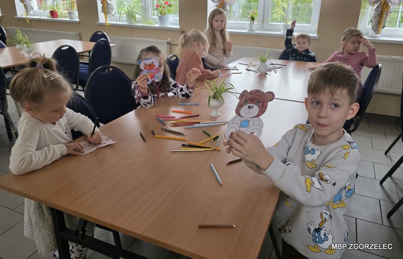 Dzieci siedzą przy stołach i kolorują papierowe misie, na stole rozłożone są kredki i wycięte elementy.