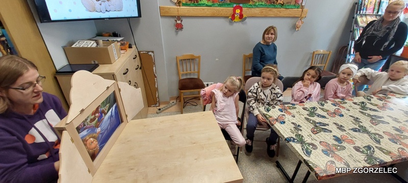 Pokaz ilustracji w teatrzyku kamishibai – dzieci siedzą przy stole, oglądając obrazki w drewnianej skrzynce.