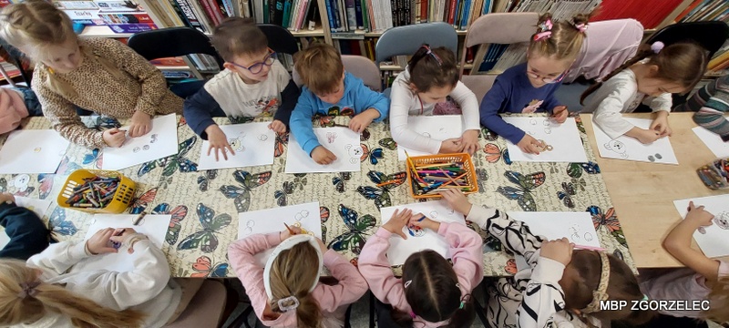 Rysowanie i kolorowanie w bibliotece – dzieci siedzą przy stole i rysują misie na kartkach papieru, na stole kredki i przybory plastyczne.