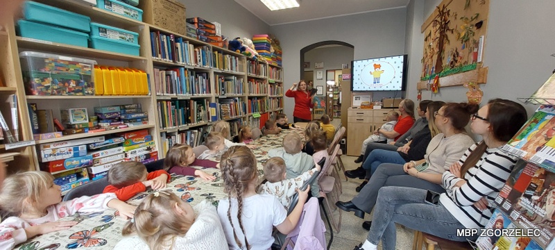 Czytanie bajek w bibliotece – dzieci siedzą przy stole, a bibliotekarka pokazuje ilustracje na ekranie, w tle regały z książkami.