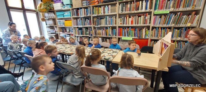 Pokaz teatrzyku kamishibai – dzieci siedzą przy stole, a bibliotekarka prezentuje ilustracje w drewnianej skrzynce kamishibai.