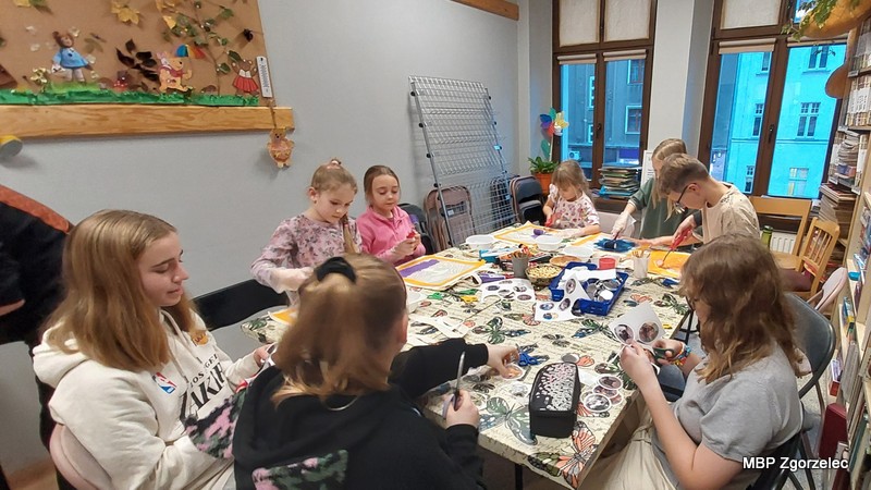 Zdjęcie przedstawia dzieci i&nbsp;młodzież pracujące przy dużym stole w&nbsp;bibliotece, podczas warsztatów plastycznych polegających na malowaniu toreb oraz tworzeniu magnesów i&nbsp;przypinek z&nbsp;motywami zwierząt.