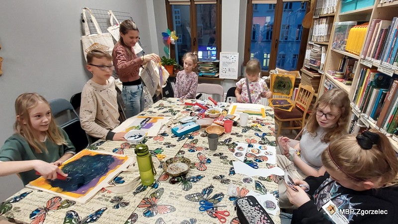 Zdjęcie przedstawia dzieci i&nbsp;młodzież pracujące przy dużym stole w&nbsp;bibliotece, podczas warsztatów plastycznych polegających na malowaniu toreb oraz tworzeniu magnesów i&nbsp;przypinek z&nbsp;motywami zwierząt.