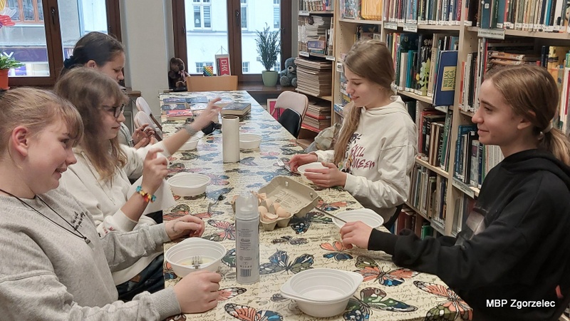 Przy długim stole bibliotecznym kilku uczestników miesza składniki w miseczkach. Na blacie stoją produkty spożywcze, w tym jajka i bita śmietana w sprayu. W tle znajdują się regały pełne książek oraz duże okna wychodzące na ulicę.