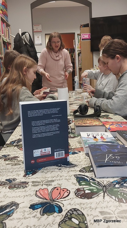 Uczestnicy Młodzieżowego Klubu Literackiego siedzą przy długim stole nakrytym obrusem z&nbsp;motylami. Na stole leżą książki, słuchawki i&nbsp;kubki, a&nbsp;w tle widać regały z&nbsp;literaturą. Osoba prowadząca stoi przy końcu stołu i&nbsp;omawia czynności związane z&nbsp;kulinarną częścią spotkania.