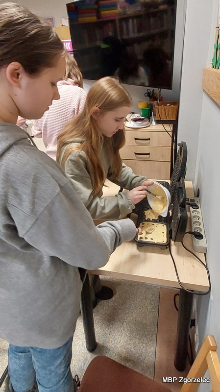 Przy stole ustawionym w&nbsp;sali bibliotecznej dwie osoby biorą udział w&nbsp;przygotowywaniu gofrów – jedna trzyma miskę z&nbsp;ciastem, a&nbsp;druga nadzoruje proces napełniania gofrownicy. W&nbsp;tle znajdują się regały i&nbsp;sprzęt wykorzystywany podczas zajęć.
