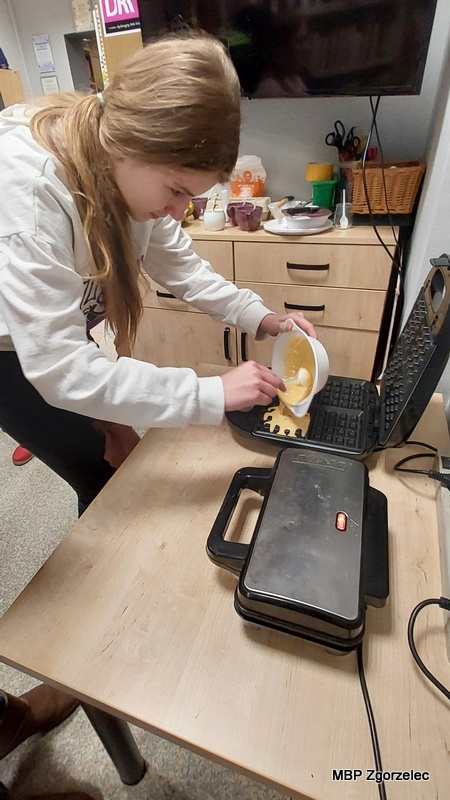 Osoba uczestnicząca w&nbsp;spotkaniu stoi przy stole kuchennym w&nbsp;bibliotece i&nbsp;przelewa porcję ciasta z&nbsp;miski do otwartej gofrownicy. W&nbsp;tle widoczne są szafki, akcesoria kuchenne oraz telewizor zawieszony na ścianie.