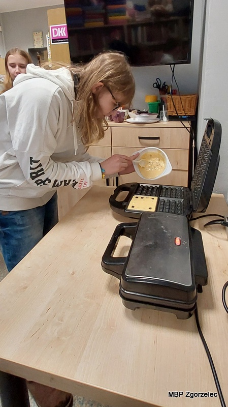 Osoba uczestnicząca w&nbsp;spotkaniu stoi przy stole kuchennym w&nbsp;bibliotece i&nbsp;przelewa porcję ciasta z&nbsp;miski do otwartej gofrownicy. W&nbsp;tle widoczne są szafki, akcesoria kuchenne oraz telewizor zawieszony na ścianie.