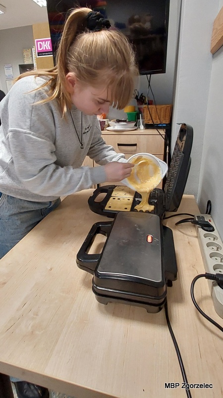 Osoba uczestnicząca w&nbsp;spotkaniu stoi przy stole kuchennym w&nbsp;bibliotece i&nbsp;przelewa porcję ciasta z&nbsp;miski do otwartej gofrownicy. W&nbsp;tle widoczne są szafki, akcesoria kuchenne oraz telewizor zawieszony na ścianie.